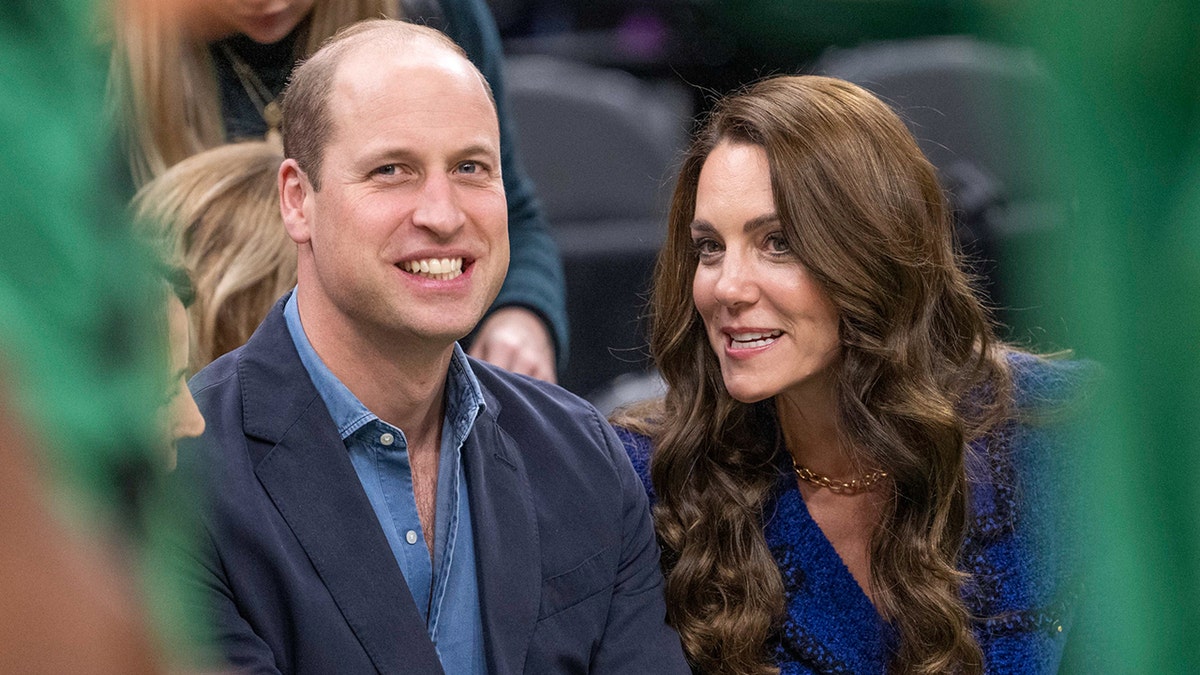 Kate Middleton in a royal blue vintage Chanel blazer leans in to her husband Prince William in discussion on the floor of the Boston Celtics game at TD Garden