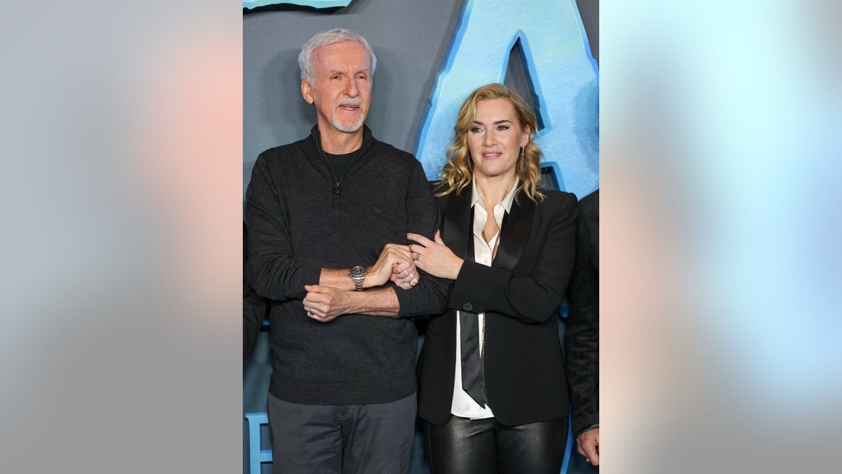 James Cameron in all black and Kate Winslet in a white top and black suit hold hands on the carpet of "Avatar: The Way of Water"