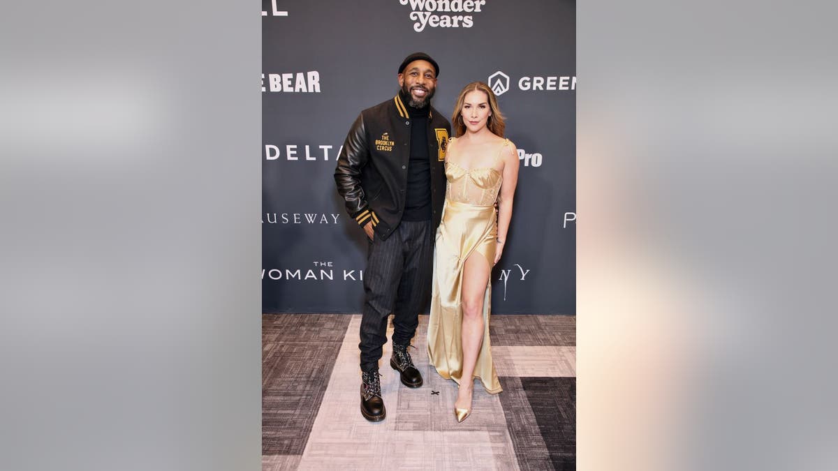Stephen "tWitch" Boss in a black bomber jacket and black pants poses with wife Allison in a shimmery gold dress