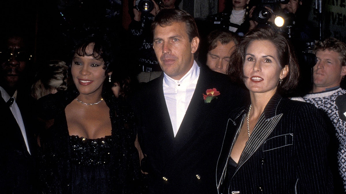Whitney Houston in a black dress poses next to Kevin Costner in a black suit and his wife Cindy in black