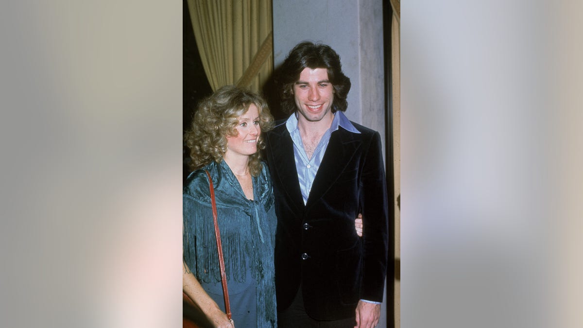 John Travolta in a velvet suit and blue undershirt with his girlfriend Dianna Hyland in a teal coat