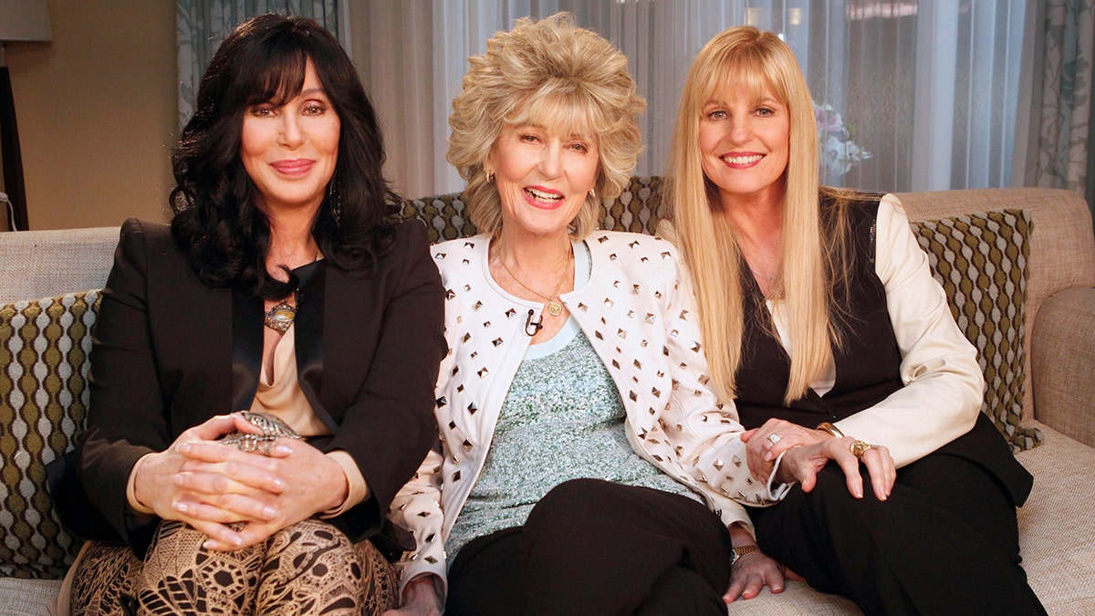 Cher smiles in a black cardigan and tan dress with her mother in the middle in a blue shirt and white and red cardigan next to her daughter Georganne LaPiere in black