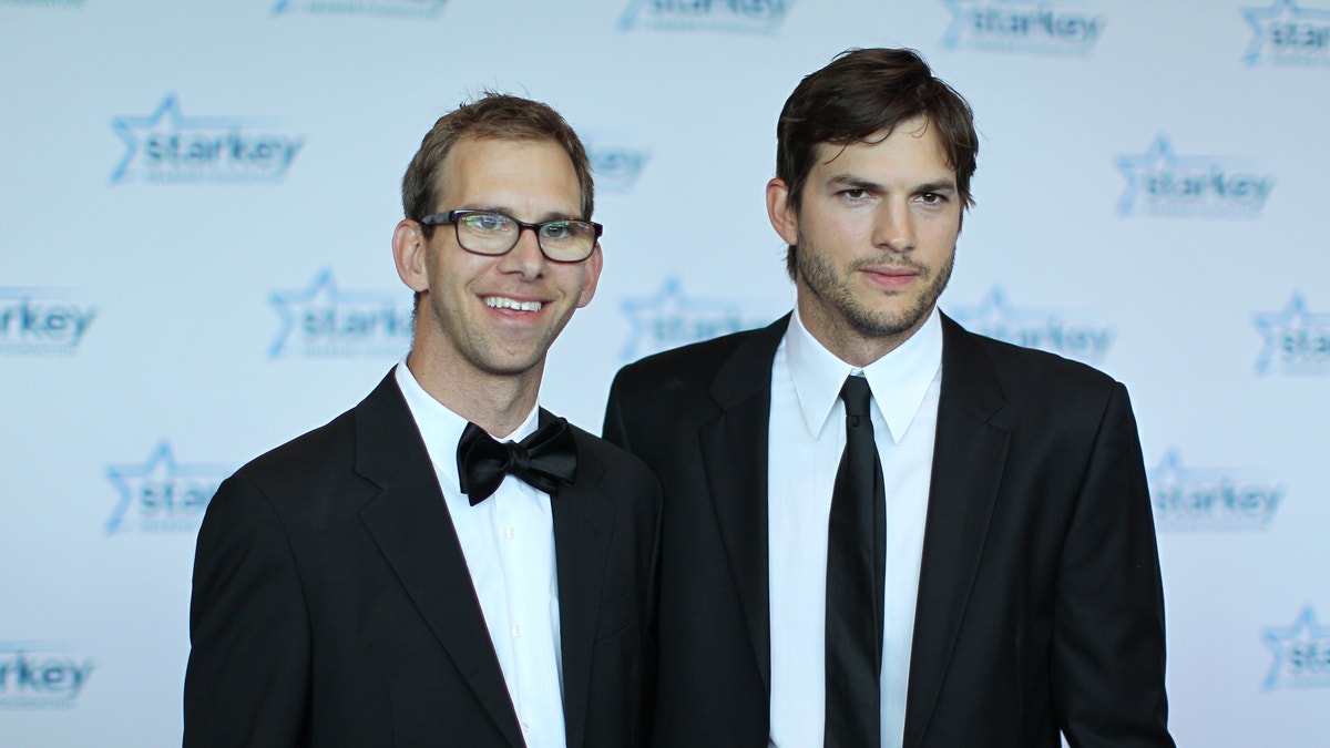 Michael Kutcher in a black tuxedo smiles next to his twin brother Ashton Kutcher in a black suit and tie