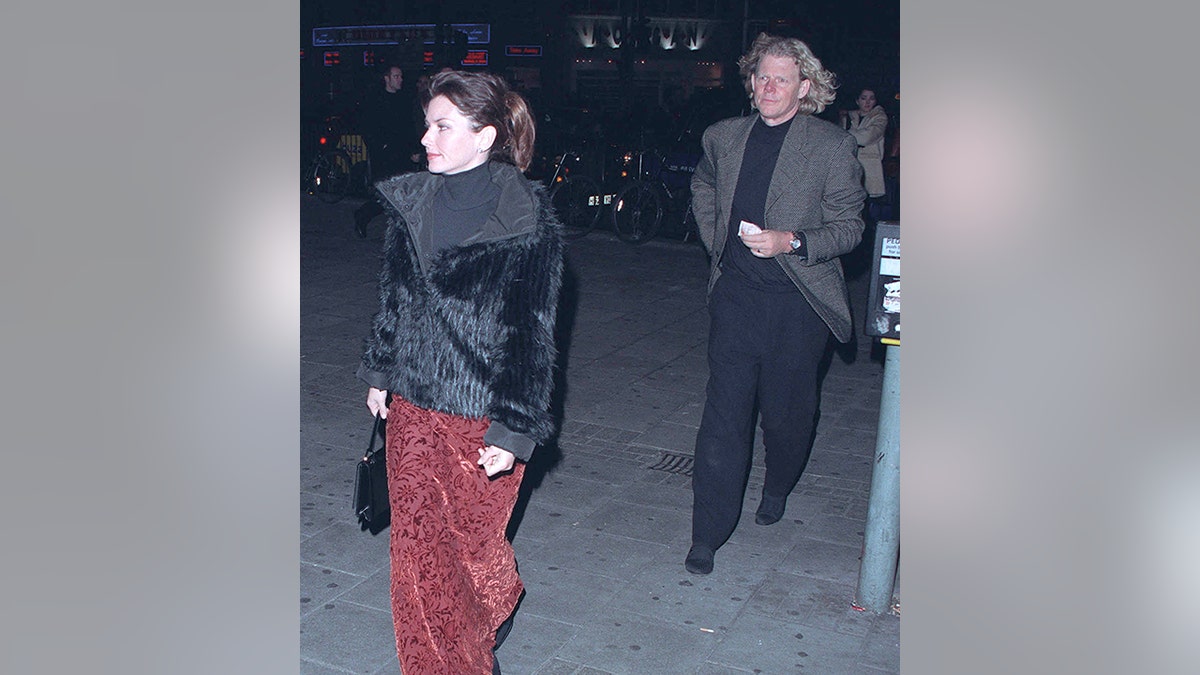 Shania Twain in a red patterned velvet skirt, a black top and black jacket walks ahead of husband Robert John Mutt Lange in black pants and a grey jacket