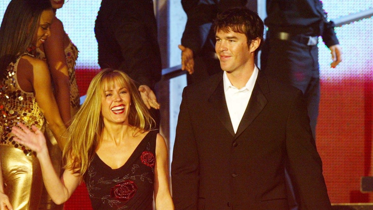 Trista in a black dress with red flowers holds the hand of Ryan Sutter in a black suit