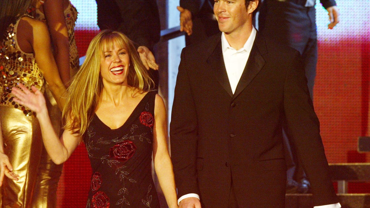 Trista in a black dress with red flowers holds the hand of Ryan Sutter in a black suit