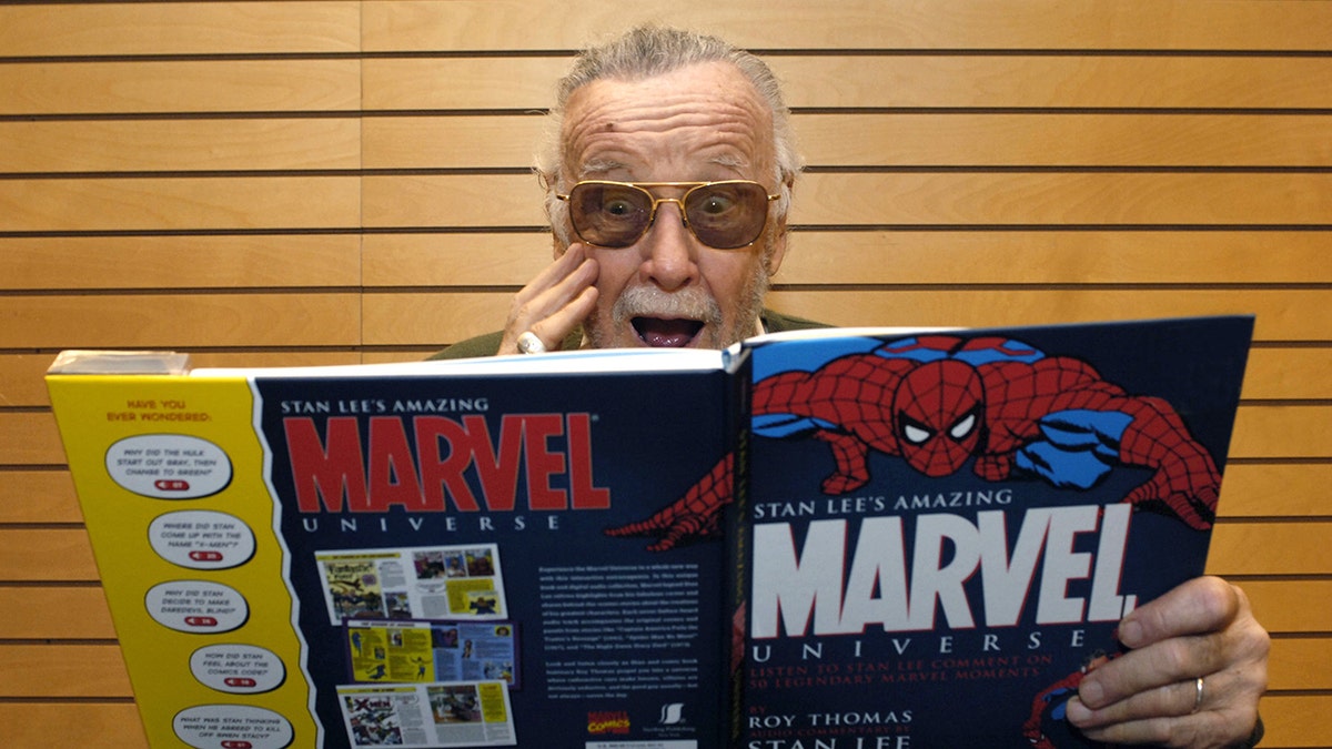 Stan Lee puts his hand to his face in a shocked expression while reading a massive Marvel Comic book
