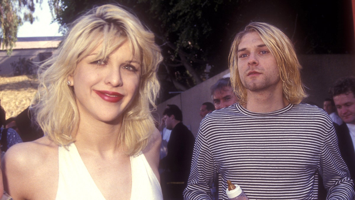Courtney Love in white smiles at the camera while Kurt Cobain in a striped shirt is photographed in the background