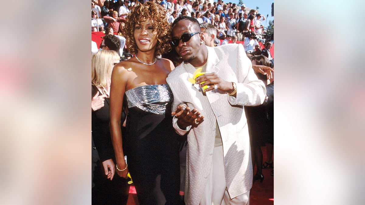 Bobby Brown in an all-white suit stands next to Whitney Houston in a black dress with a sparkly top at the Emmy Awards