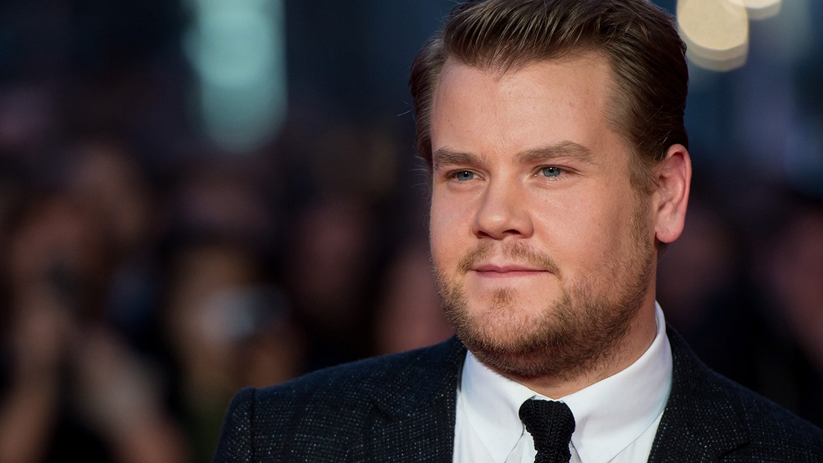 James Corden in a black suit, white shirt, and black skinny tie looks off in the distance on the red-carpet in London