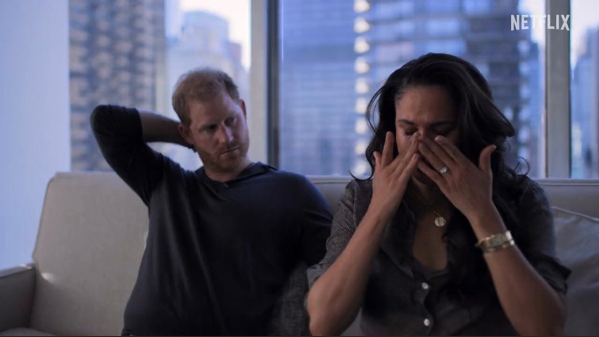 Meghan Markle cries on the sofa with her hands over her face as Prince Harry looks on with his arm behind his neck