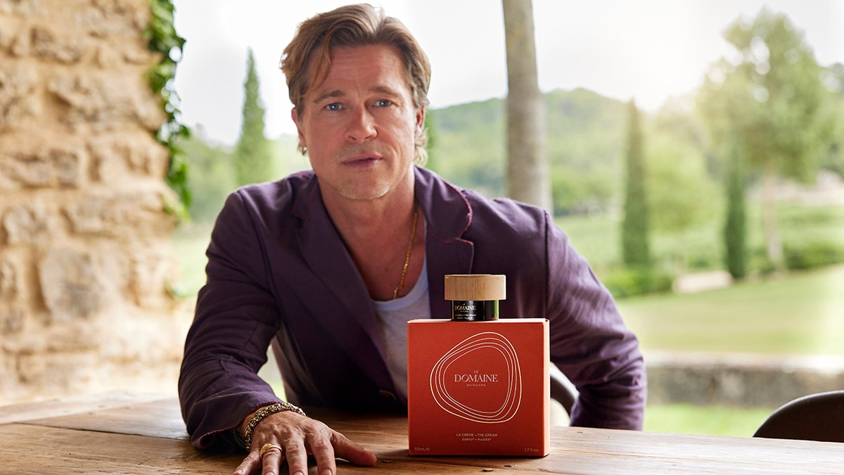 Brad Pitt launches skincare line in a photo wearing a purple shirt and a large red box of his new product