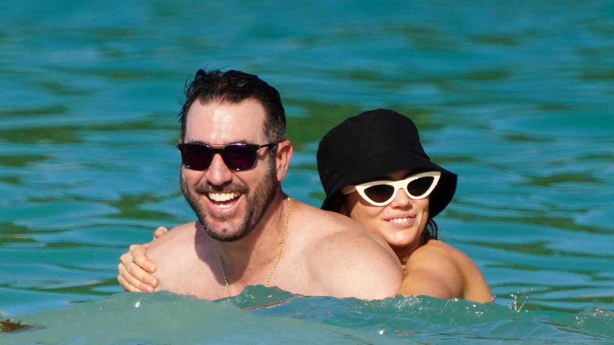 Kate Upton in a black bucket hat and white cat-eye sunglasses holds on to her husband Justin Verlander in the water