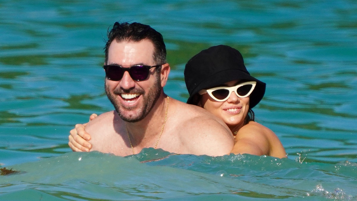 Kate Upton in a black bucket hat and white cat-eye sunglasses holds on to her husband Justin Verlander in the water