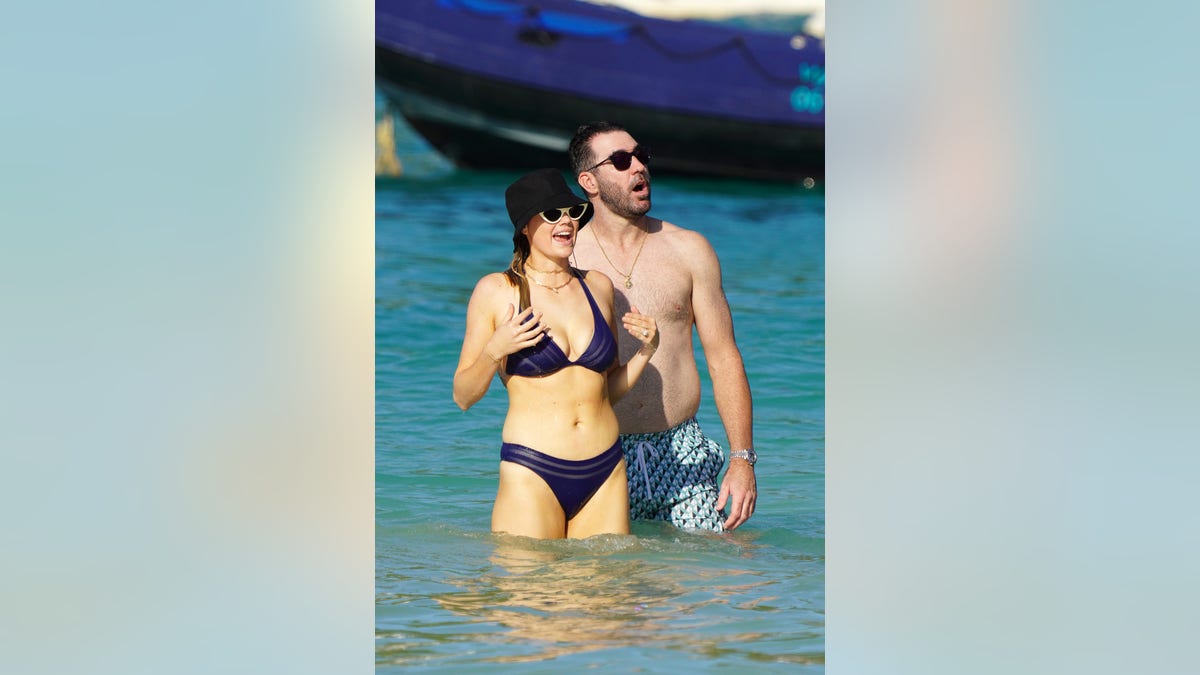 Kate Upton in a blue bikini and black bucket hat and white cat-eye sunglasses looks shocked at something next to her husband Justin Verlander in a turquoise patterned wimsuit