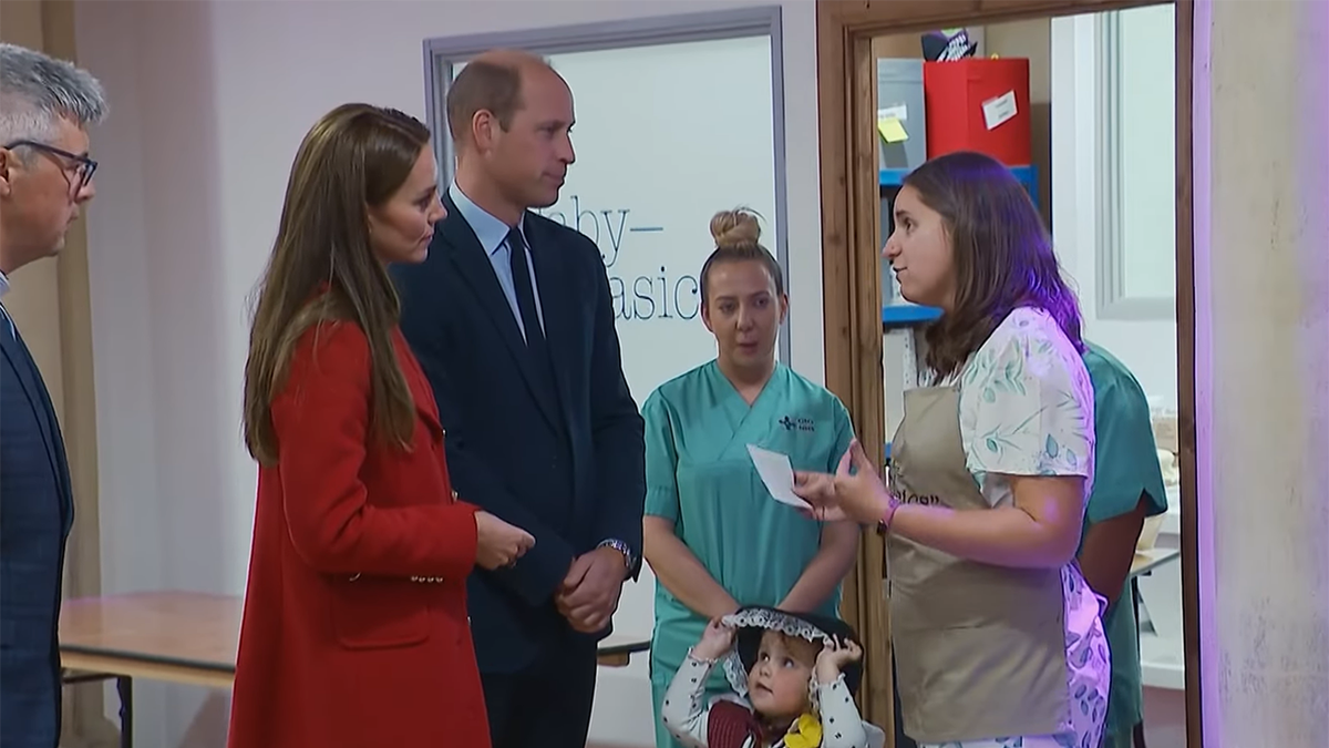 Kate Middleton in a red coat and Prince William in a blue suit talk to medical professionalsi