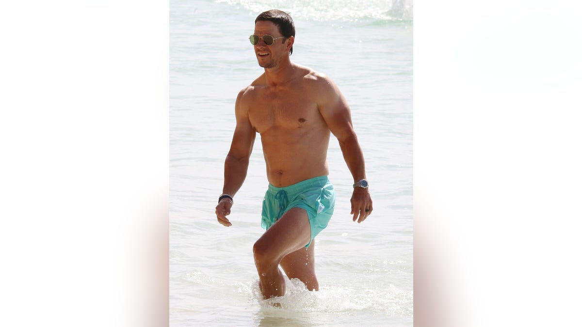 Mark Wahlberg walking out of the water with turquoise swim trunks, wearing his sunglasses