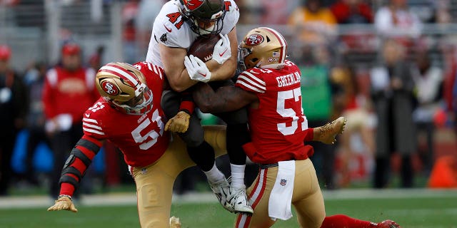 El ala cerrada de los Tampa Bay Buccaneers, Ko Kieft, en el centro, es abordado por el apoyador de los 49ers de San Francisco, Azeez Al-Shaair (51), y el apoyador, Dre Greenlaw (57), durante la primera mitad de un juego del Campeonato de Fútbol de la NFL en Santa Clara, California, el domingo 11 de diciembre de 2022.