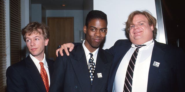 David Spade in a suit and red tie looks off camera while posing with Chris Rock in a dark suit and tie who is hugged by Chris Farley