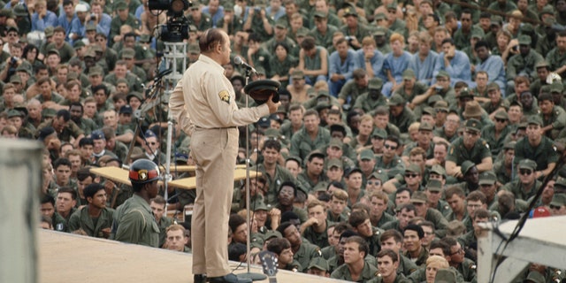 Comedian Bob Hope stands at a microphone on stage as crowds of United States troops watch the Christmas show he put on for troops at the Camp Eagle army base, southeast of Hue, Vietnam, on Dec. 22, 1970.