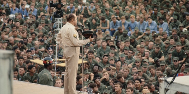 Comedian Bob Hope stands at a microphone on stage as crowds of United States troops watch the Christmas show he put on for troops at the Camp Eagle army base, southeast of Hue, Vietnam, on Dec. 22, 1970.