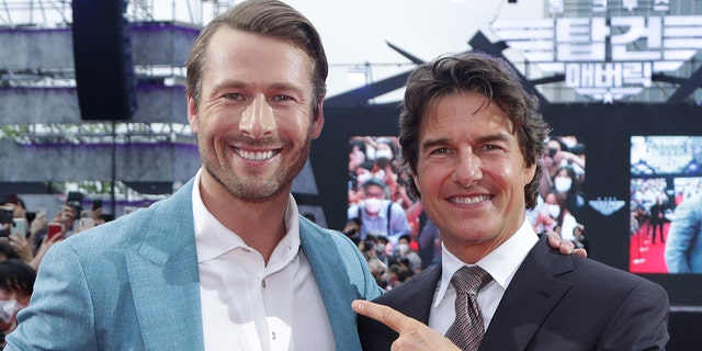 Glen Powell wearing a white button down and blue/teal blazer poses next to a pointing Tom Cruise in a black suit on the red carpet