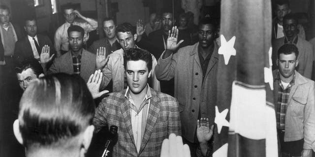 American rock 'n' roll singer Elvis Presley (1935-1977) stands with a group of young men at an induction center, raising their right hands as they are sworn into the United States Army by an officer standing next to an American flag. 