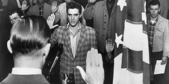 American rock 'n' roll singer Elvis Presley (1935-1977) stands with a group of young men at an induction center, raising their right hands as they are sworn into the United States Army by an officer standing next to an American flag. 