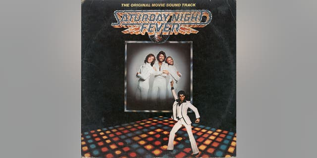 View of the cover of the soundtrack album from the film "Saturday Night Fever," 1977. Published by RSO Records, the album features a large, framed portrait of Australian pop group the Bee Gees as they overlook a photo of American actor John Travolta as he strikes a pose on a disco dance floor.