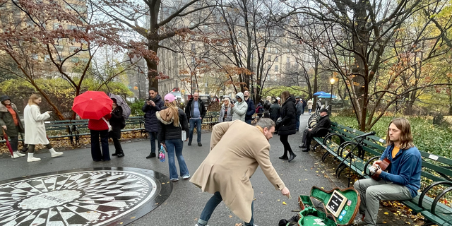 New York City musician Jules Avalon (right, on bench) performs acoustic versions of Beatles' and John Lennon's songs in Strawberry Fields, Central Park, on Dec. 7, 2022. The Dakota, the luxury residence where Lennon was murdered on Dec. 8, 1980, can be seen in the background.