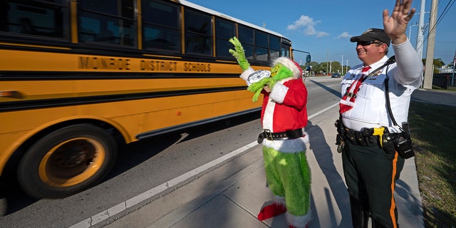 In this photo provided by the Florida Keys News Bureau, Monroe County Sheriff's Office Colonel Lou Caputo, left, costumed as the Grinch, and Deputy Andrew Leird, right, wave at a school bus rolling on the Florida Keys Overseas Highway Tuesday, Dec. 13, 2022, in Marathon, Fla. When drivers are pulled over for slightly speeding through a school zone, Caputo offers them the choice between an onion or a traffic citation. It's a holiday tradition in the Keys that Caputo began 20 years ago. 
