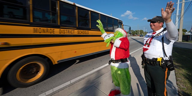 In this photo provided by the Florida Keys News Bureau, Monroe County Sheriff's Office Colonel Lou Caputo, left, costumed as the Grinch, and Deputy Andrew Leird, right, wave at a school bus rolling on the Florida Keys Overseas Highway Tuesday, Dec. 13, 2022, in Marathon, Florida When drivers are pulled over for speeding in a school zone, Caputo offers them the choice between an onion or a traffic citation. It's a holiday tradition in the Keys that Caputo began 20 years ago.