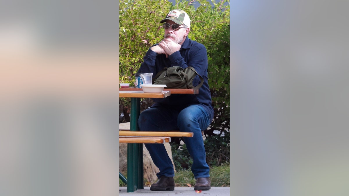 A man in a trucker hat and sunglasses looks on while seated on a bench.