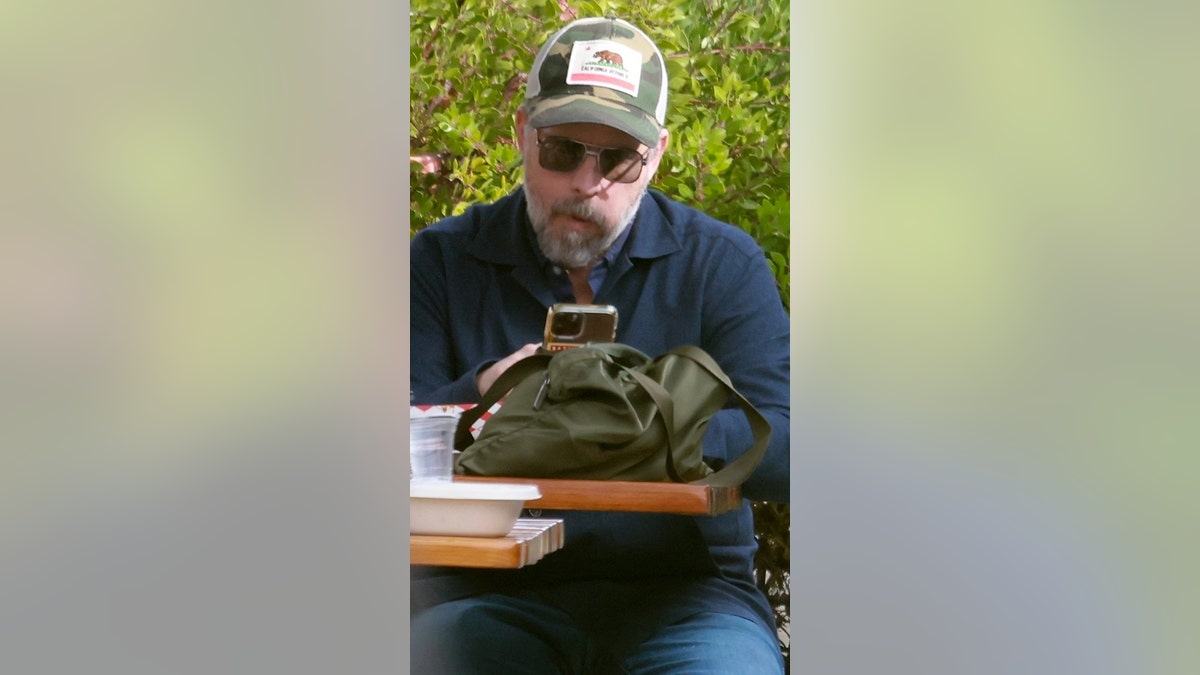 A man in sunglasses and a trucker hat sits on a bench, reading his phone.