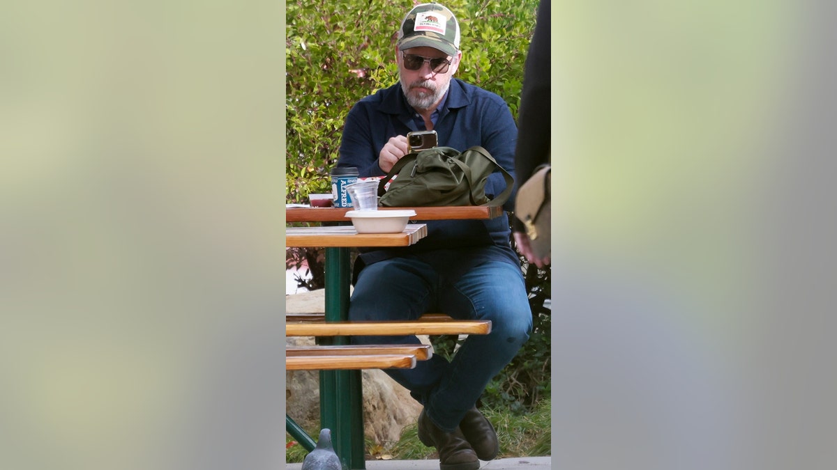 A man in sunglasses and a trucker hat sits on a bench, reading his phone.