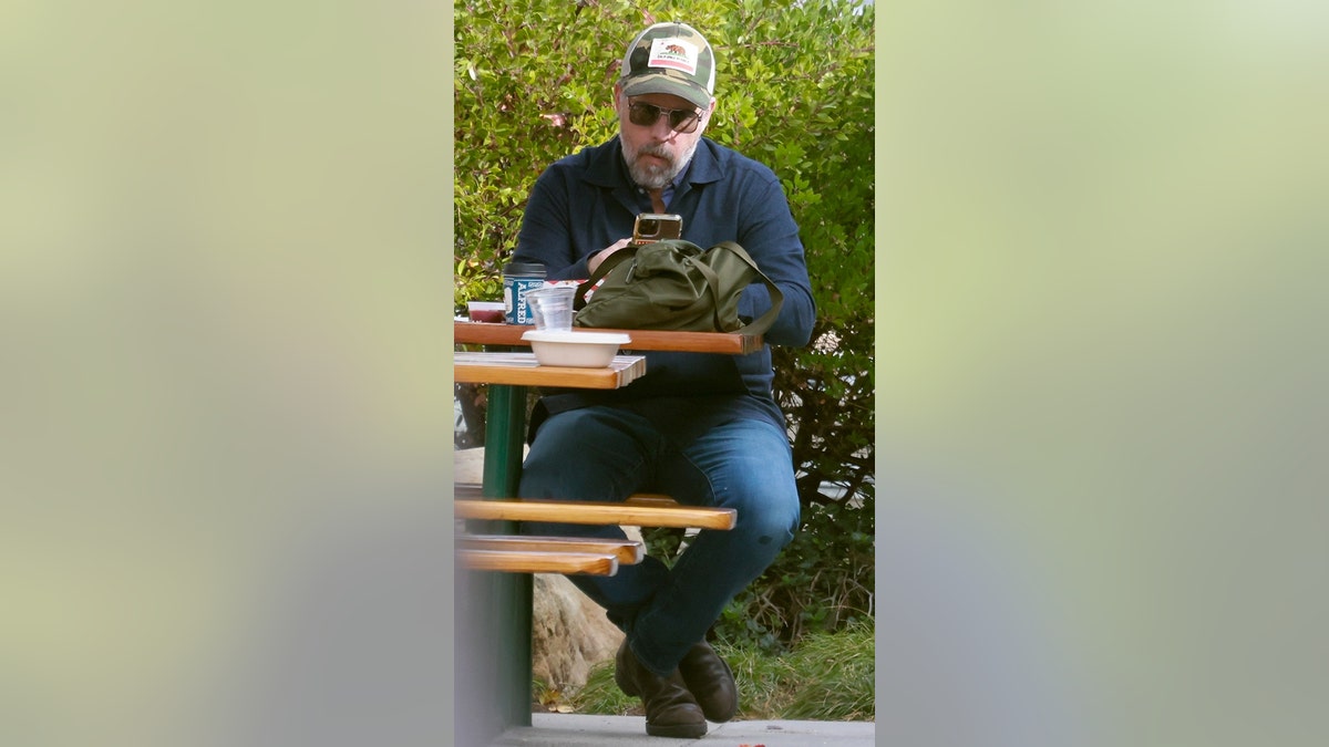 A man in sunglasses and a trucker hat sits on a bench, reading his phone.