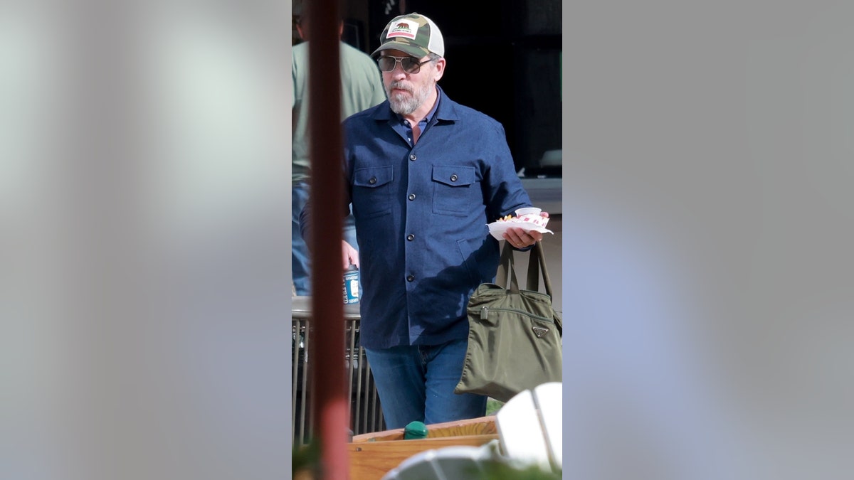 A man in a trucker hat and sunglasses carries french fries and a drink.