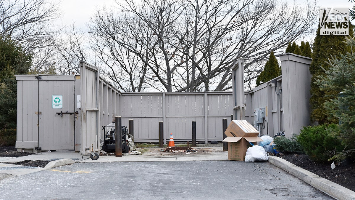 A grey wooden fence surrounds an empty dumpster area outside of an apartment complex.