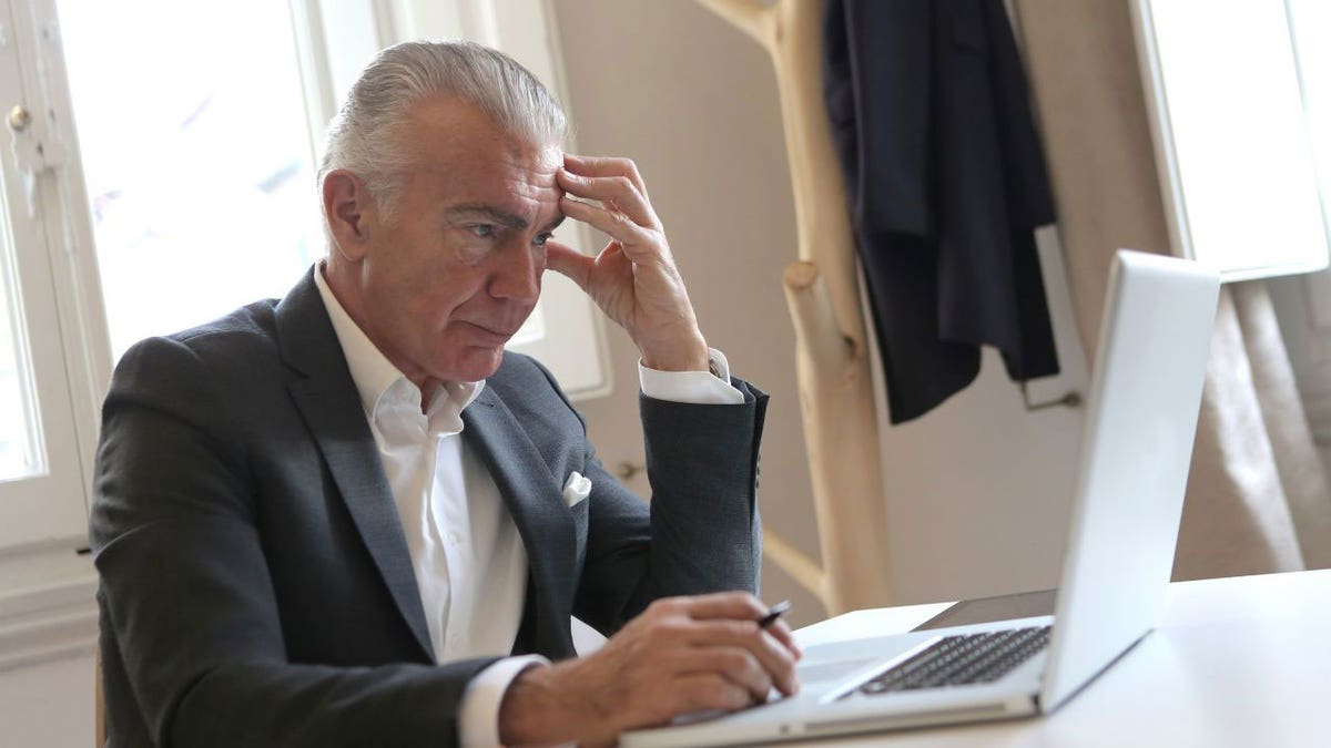 Man wearing a suit, sitting at the table, upset at his laptop.
