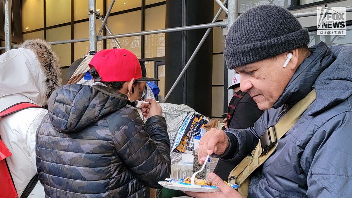 A line of people wait for food, served buffet-style on a sidewalk.