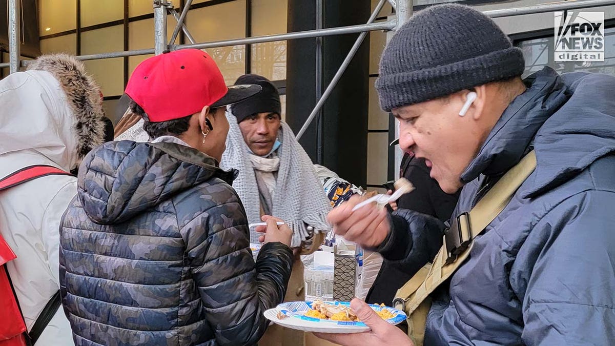 A line of people wait for food, served buffet-style on a sidewalk.