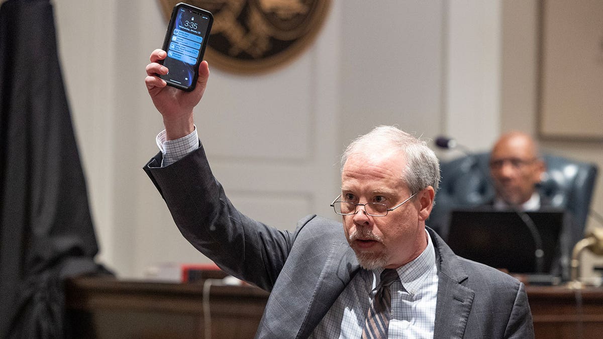 A man wearing a grey suit holds up a cell phone in court.