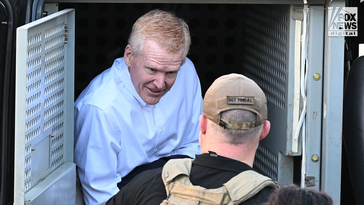 A man wearing a white button-down shirt exits a police vehicle surrounded by armed officers.