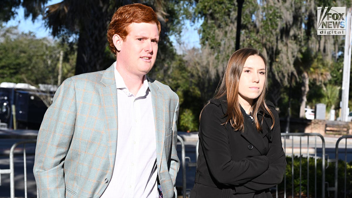 A man with red hair, wearing a grey suit enters a courthouse with a woman with brown hair.