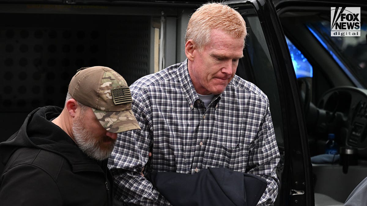 A man wearing a plaid shirt is escorted out of a police vehicle by armed officers.