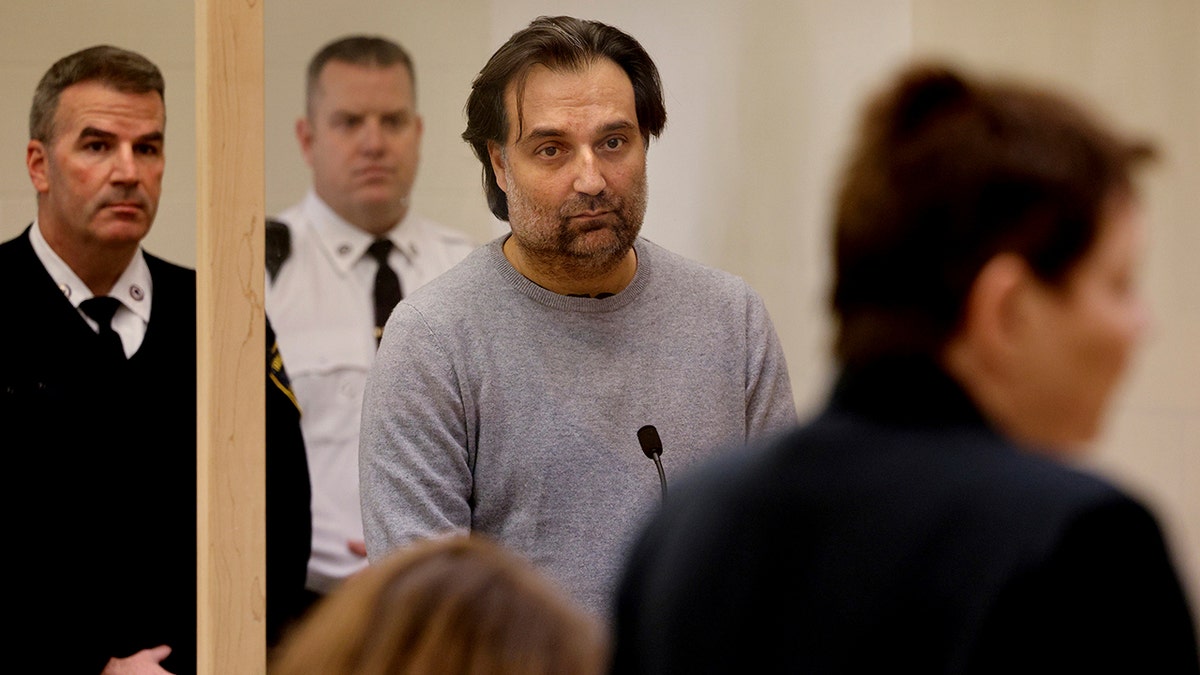A man in a grey sweatshirt stands inside of a courtroom.
