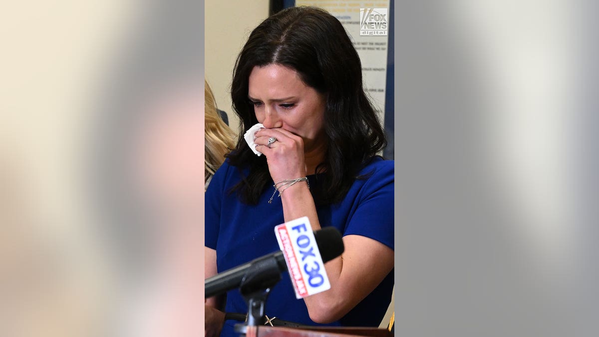 A woman wearing a blue dress cries while standing behind a press podium.