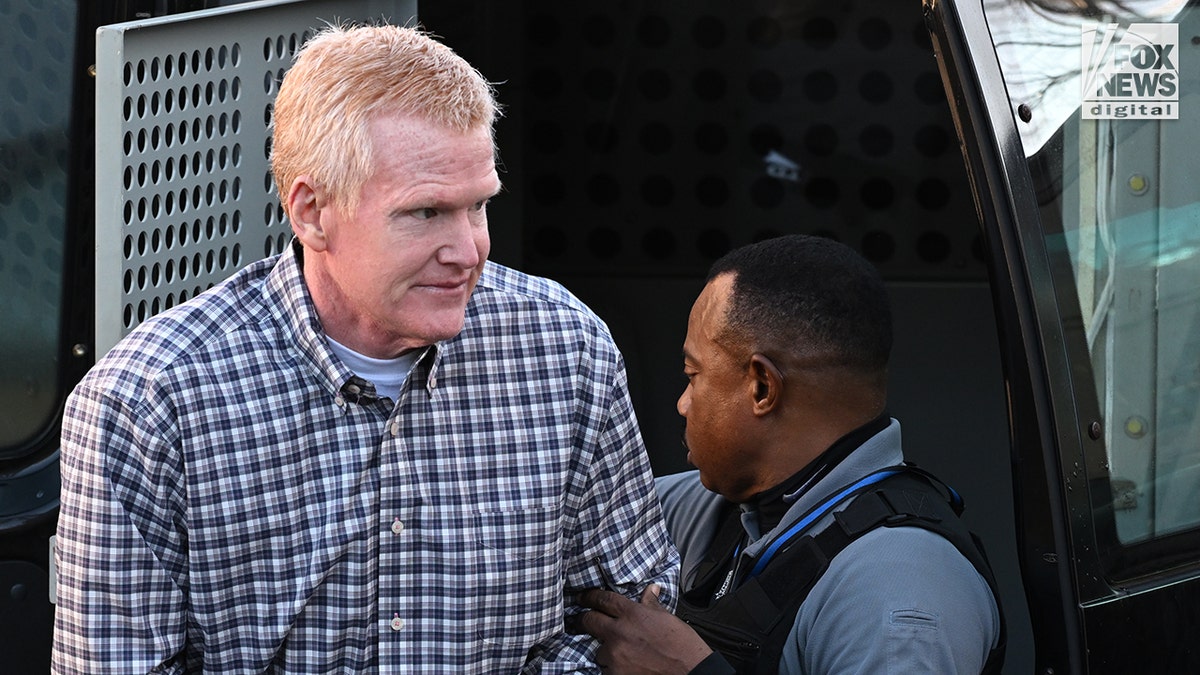 A man wearing a plaid shirt is escorted out of a black van by police.