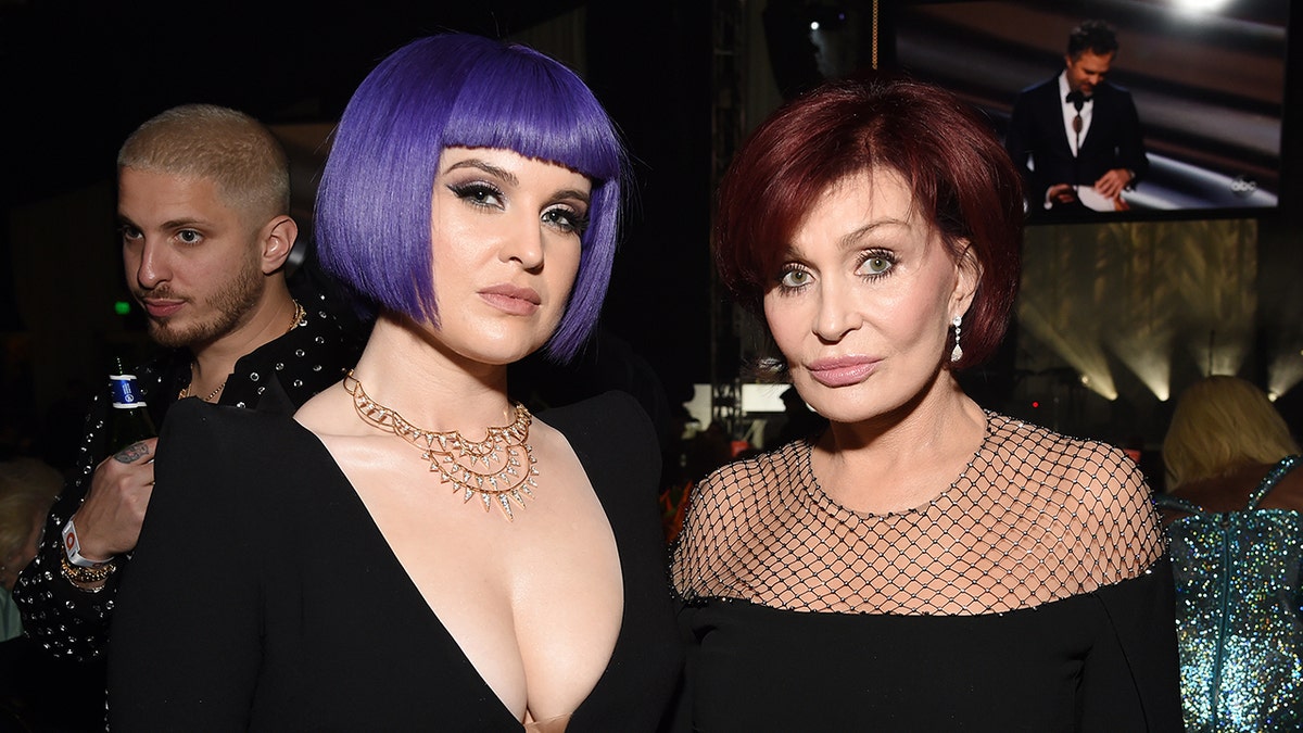 Kelly Osbourne with a short purple bob and deep V-neck black dress smiles for a photo with her mother Sharon in a black gown with a mesh top