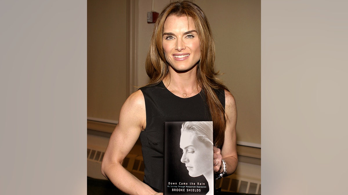 Brooke Shields in a sleeveless black outfit holds her book "Down Came the Rain: My Journey Through Postpartum Depression"